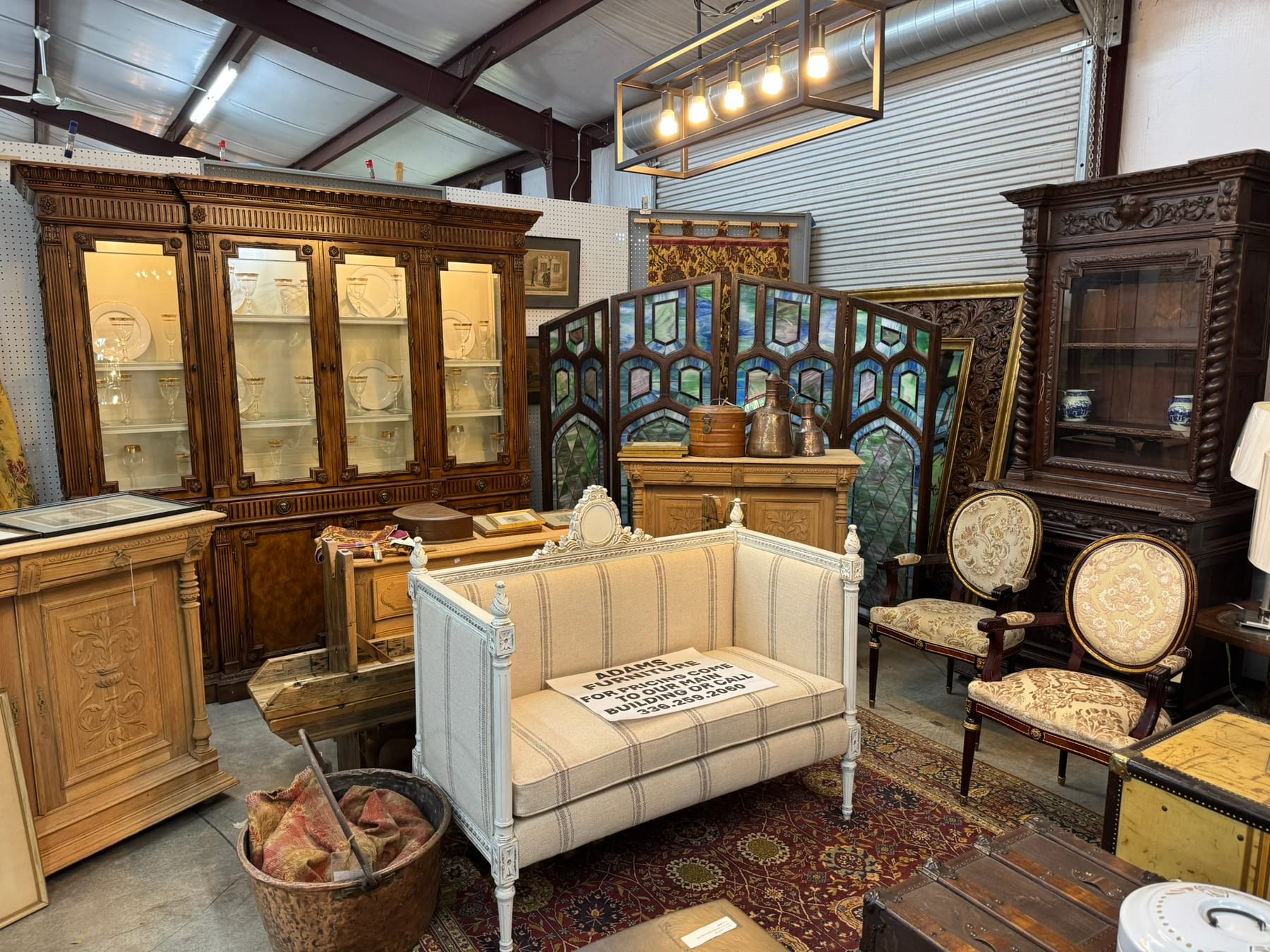 The antiques side of the booth — a carved breakfront cabinet with gilt-rimmed glassware, a stained-glass screen, a striped French settee, and a pair of oval-back armchairs