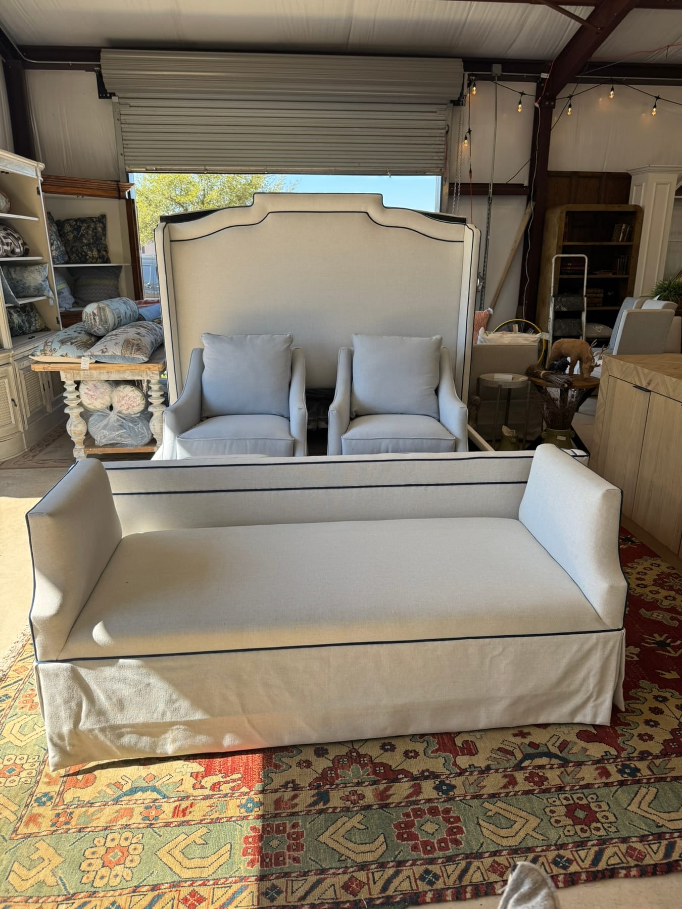 A tall upholstered headboard with navy piping, a pair of pale blue armchairs, and a matching skirted bench on a red kilim rug
