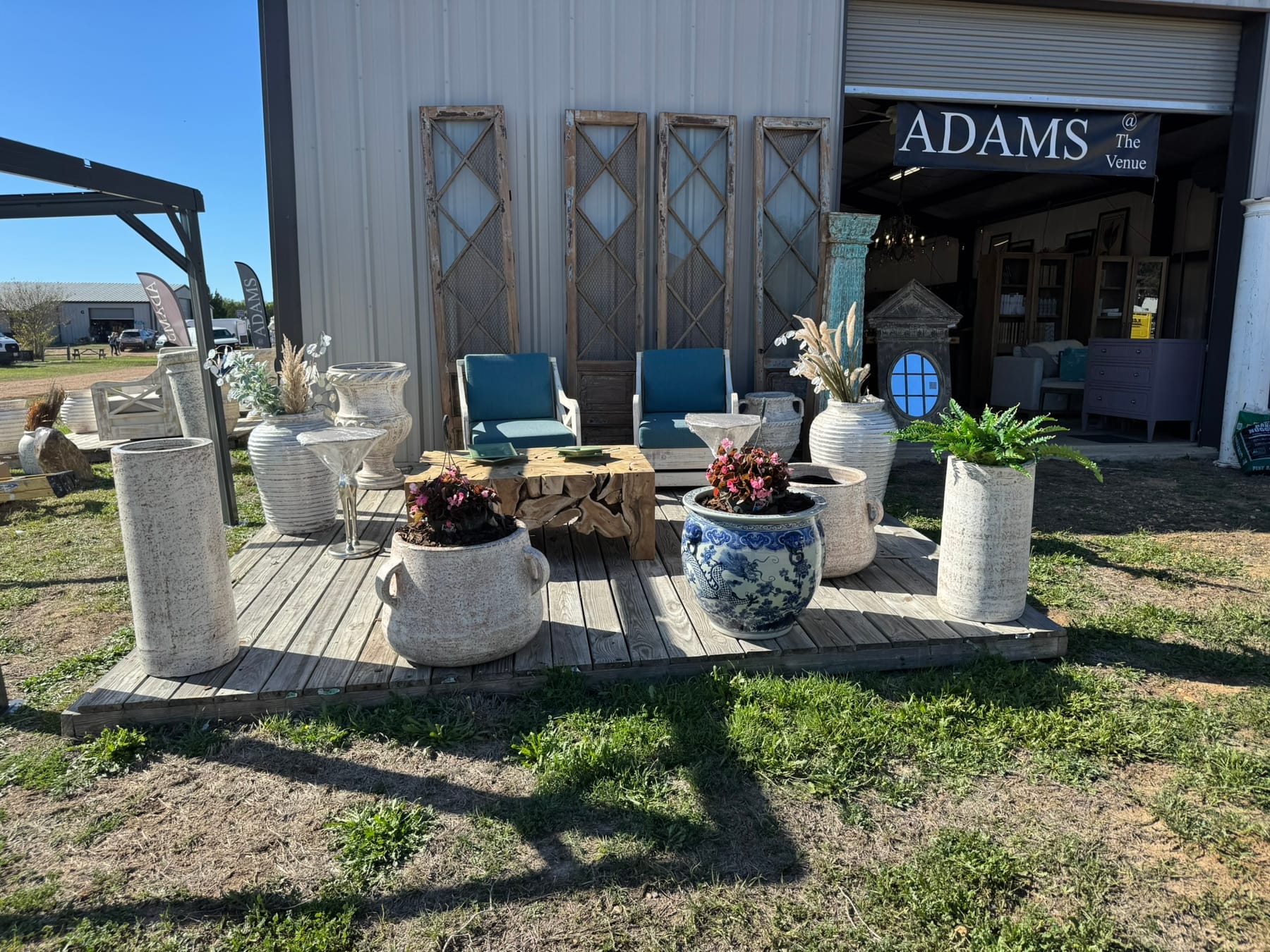 The Adams Furniture booth at The Venue in Round Top, Texas — a deck of stone planters, a teak root coffee table, and blue-cushioned armchairs in front of the open bay, with the Adams @ The Venue banner overhead