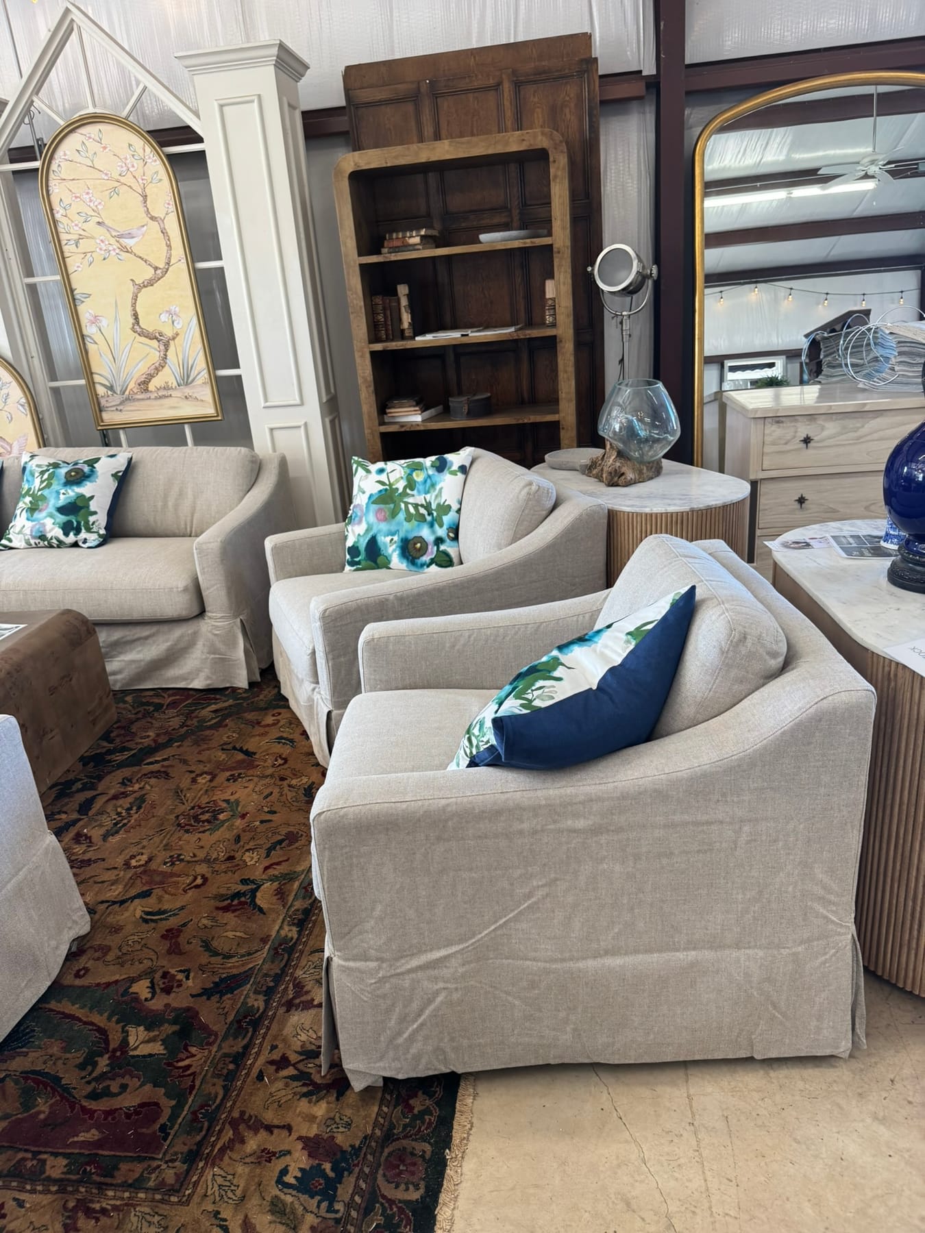 Two linen slipcovered swivel chairs with watercolor floral pillows beside a fluted marble-top side table and an arched brass mirror